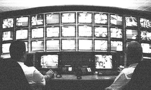 Two people sitting in front of a wall of security monitors