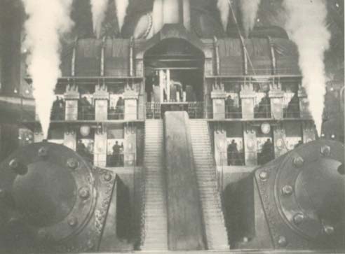 A black and white photograph of a large industrial steam engine or factory interior with steam rising