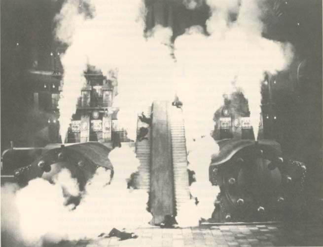 A black and white photograph of a steam locomotive emitting large clouds of steam or smoke in a station or industrial setting.